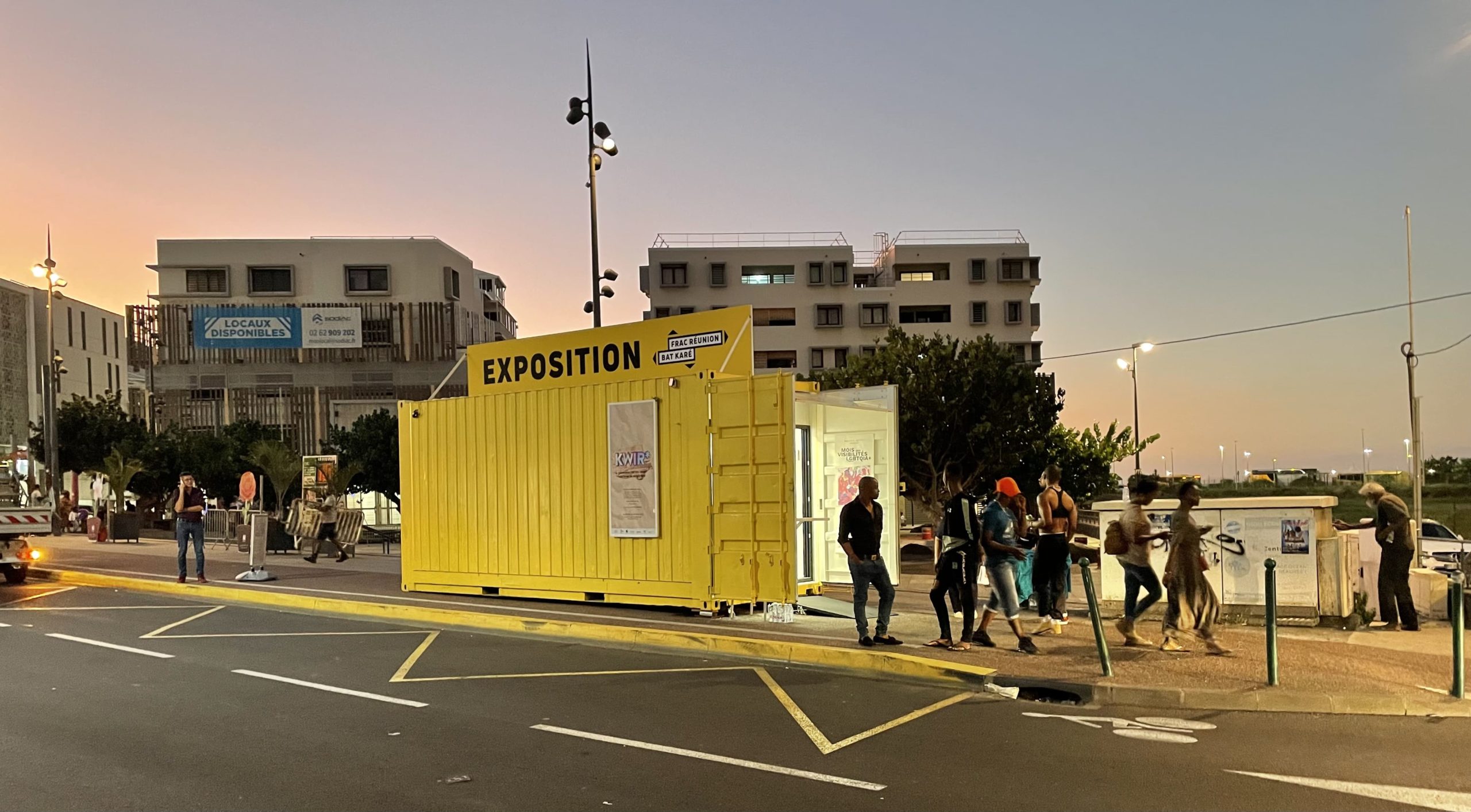 Un container jaune est situé à côté d'une route et de bâtiments. Sur son toit, il est écrit : " Exposition ". Autour, des personnes circulent. 