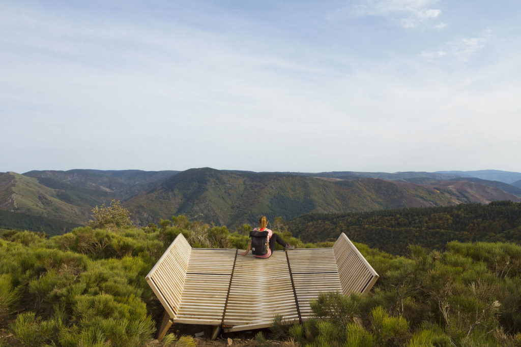 Une personne regarde un paysage assise en tailleur sur une installation en bois. 